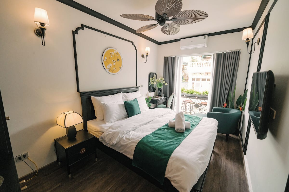 Habitación de hotel con cama, mesita de noche, lámpara, ventilador de techo y vistas al exterior.