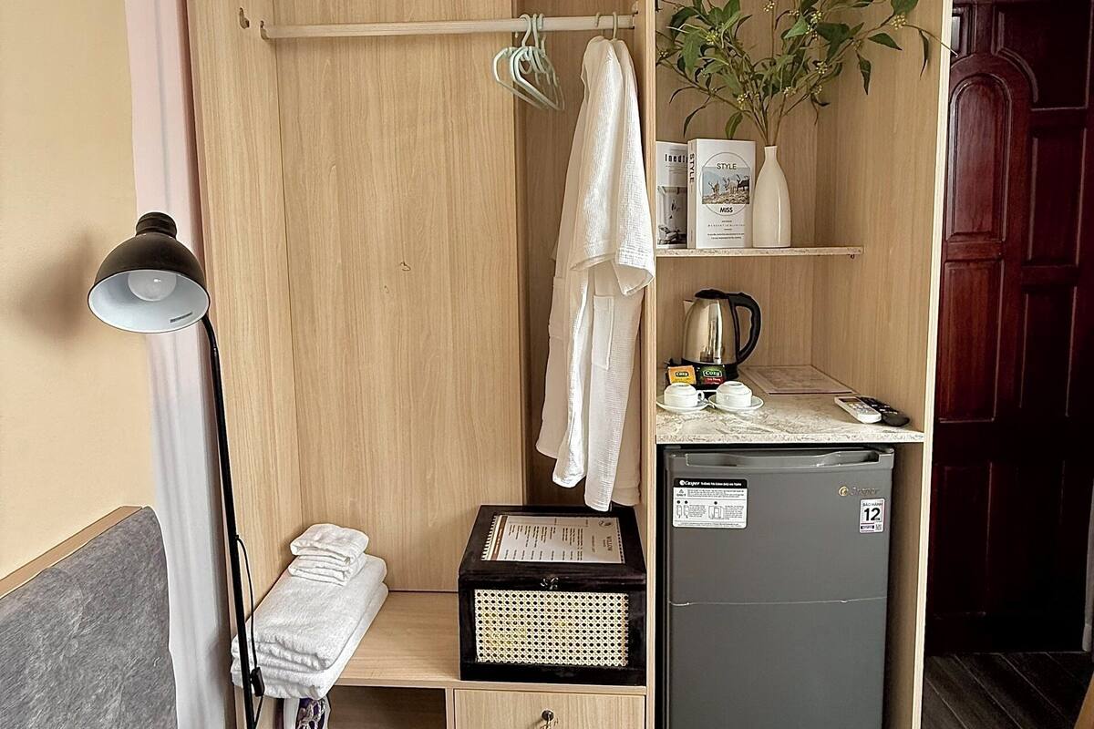 Una habitación de hotel compacta con un armario de madera, un refrigerador pequeño, una mesita de noche y una lámpara.