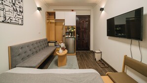 A compact hotel room with a bed, a sofa, a small wooden table, a television, and a kitchenette.