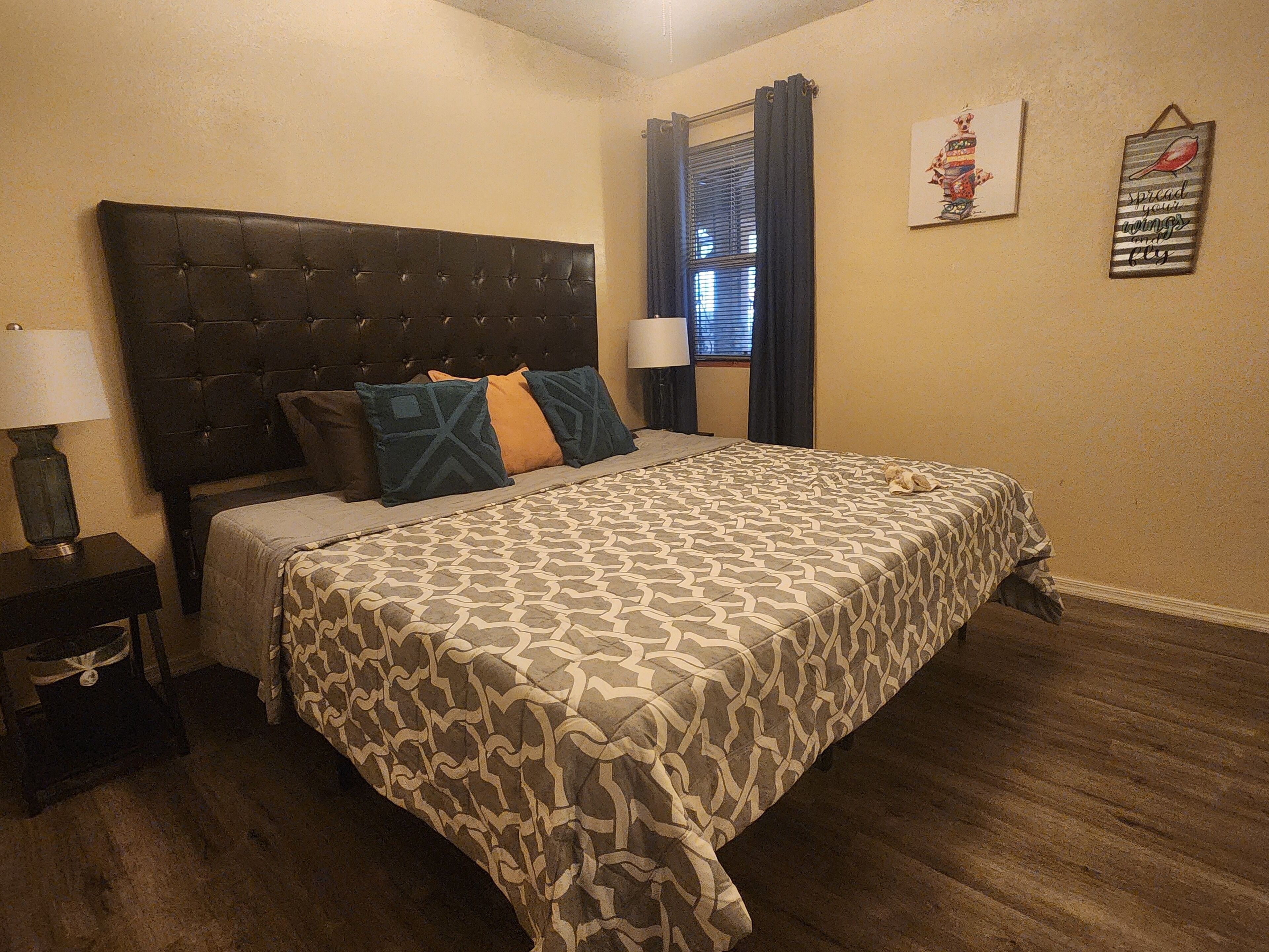 A bedroom with a tufted headboard, patterned bedspread, a nightstand with a lamp, a window with curtains, and wall art.
