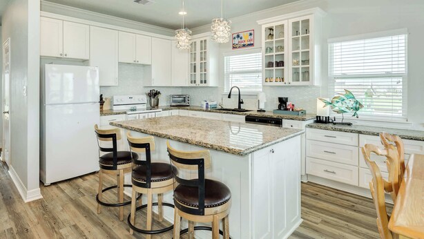 A modern kitchen with white cabinetry, a granite island, and bar stools. - Purple Paradise- Beachy Fun, Bay & Beach Views! (Surfside Beach)