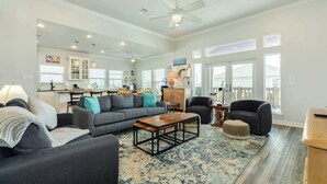 A spacious living room with a gray sofa, a coffee table, and a dark armchair. There is a kitchen in the background with white cabinets and a dining area. - Purple Paradise- Beachy Fun, Bay & Beach Views! (Surfside Beach)