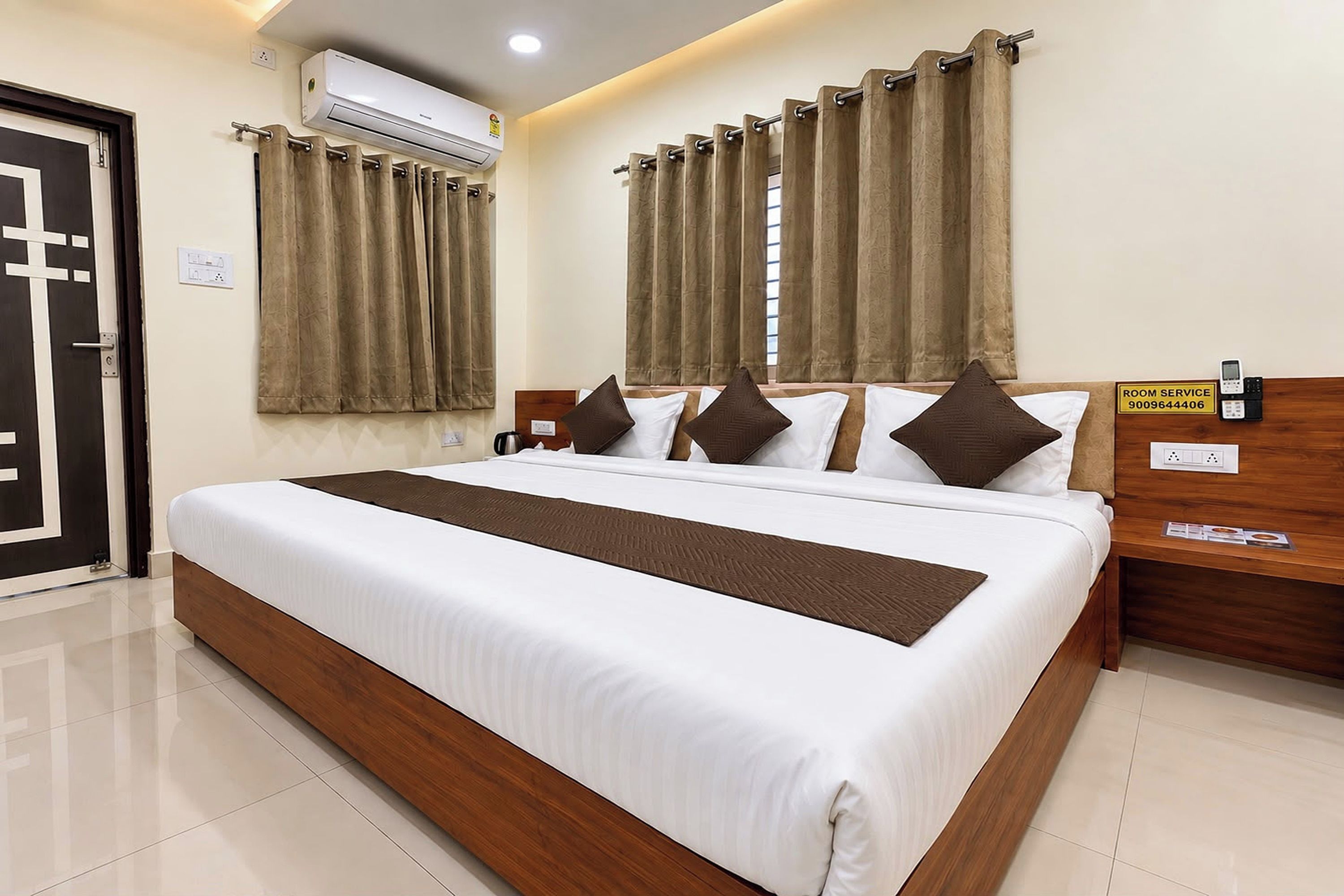 Une chambre d’hôtel avec un grand lit, des rideaux, un appareil de climatisation, une table de chevet avec un téléphone et un panneau indiquant le service en chambre.