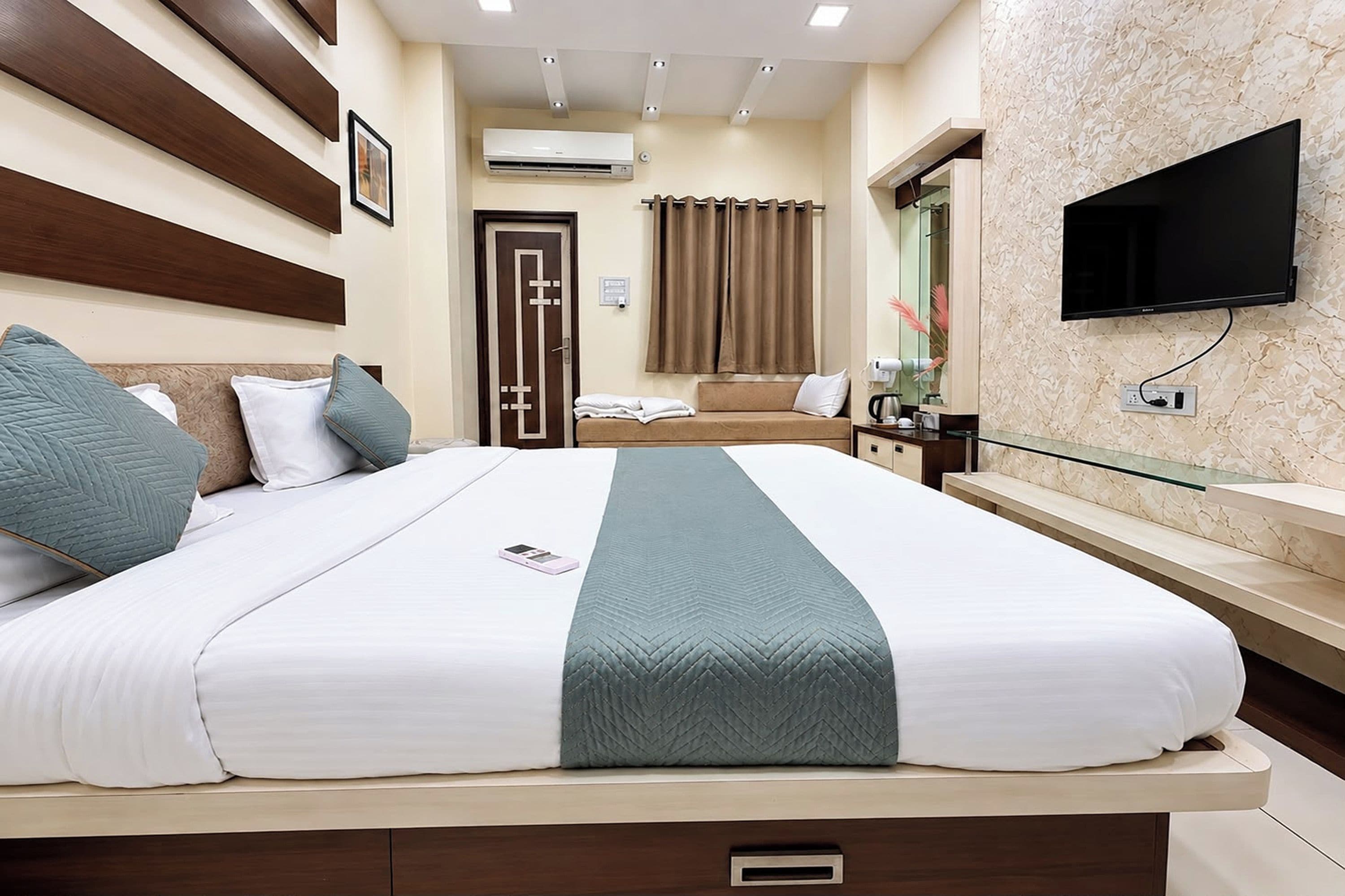 A hotel room with a bed, a bedside table, a TV, and a bathroom visible in the background.