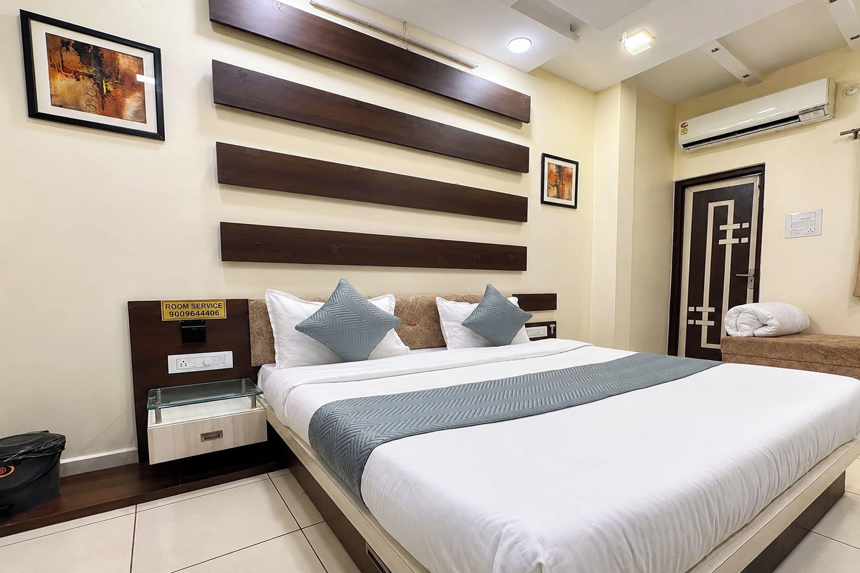 A hotel room with a bed, a bedside table, a nightstand, a wall-mounted air conditioning unit, and a door.