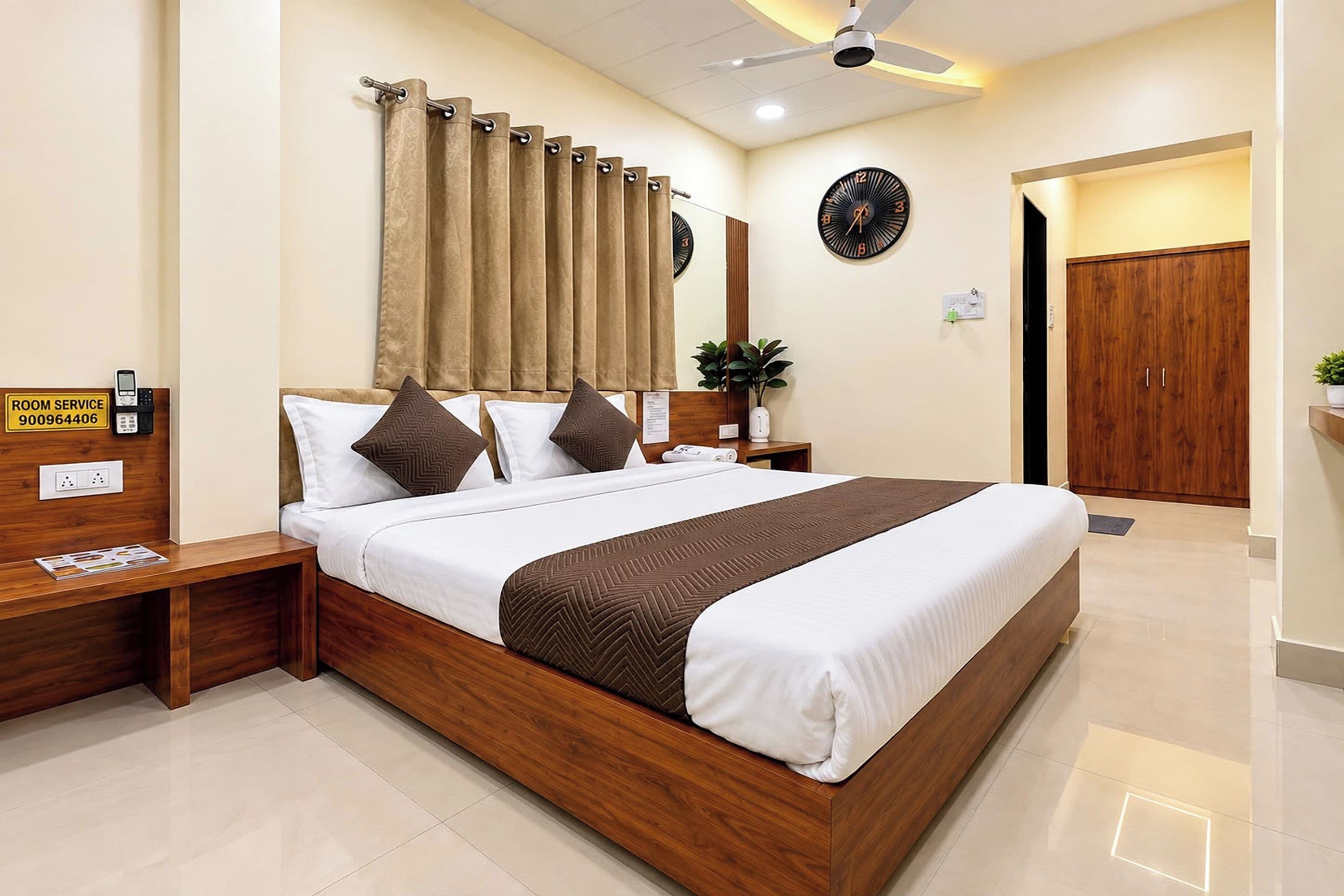 A hotel room with a wooden bed, a bedside table, a nightstand, a telephone, a clock, a mirror, a door, and a plant.