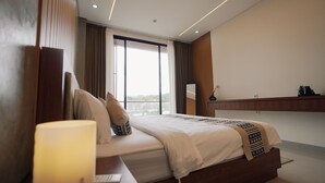 A modern hotel room with a large bed, a bedside table, a nightstand, a window with curtains, and a wall-mounted TV. - SLOKAYANA VILLAS (Pecatu)