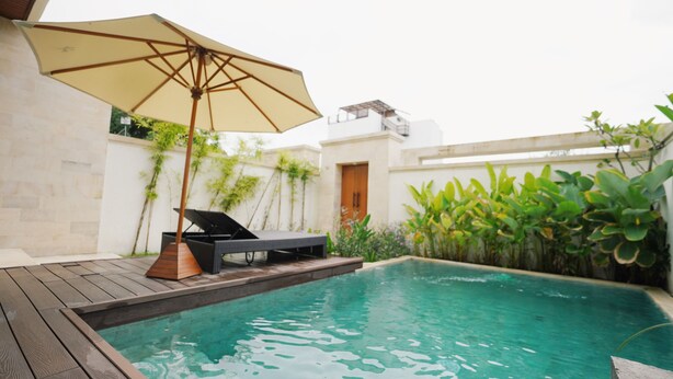 A swimming pool with a wooden deck and lounge chairs, surrounded by greenery and a white wall. - SLOKAYANA VILLAS (Pecatu)