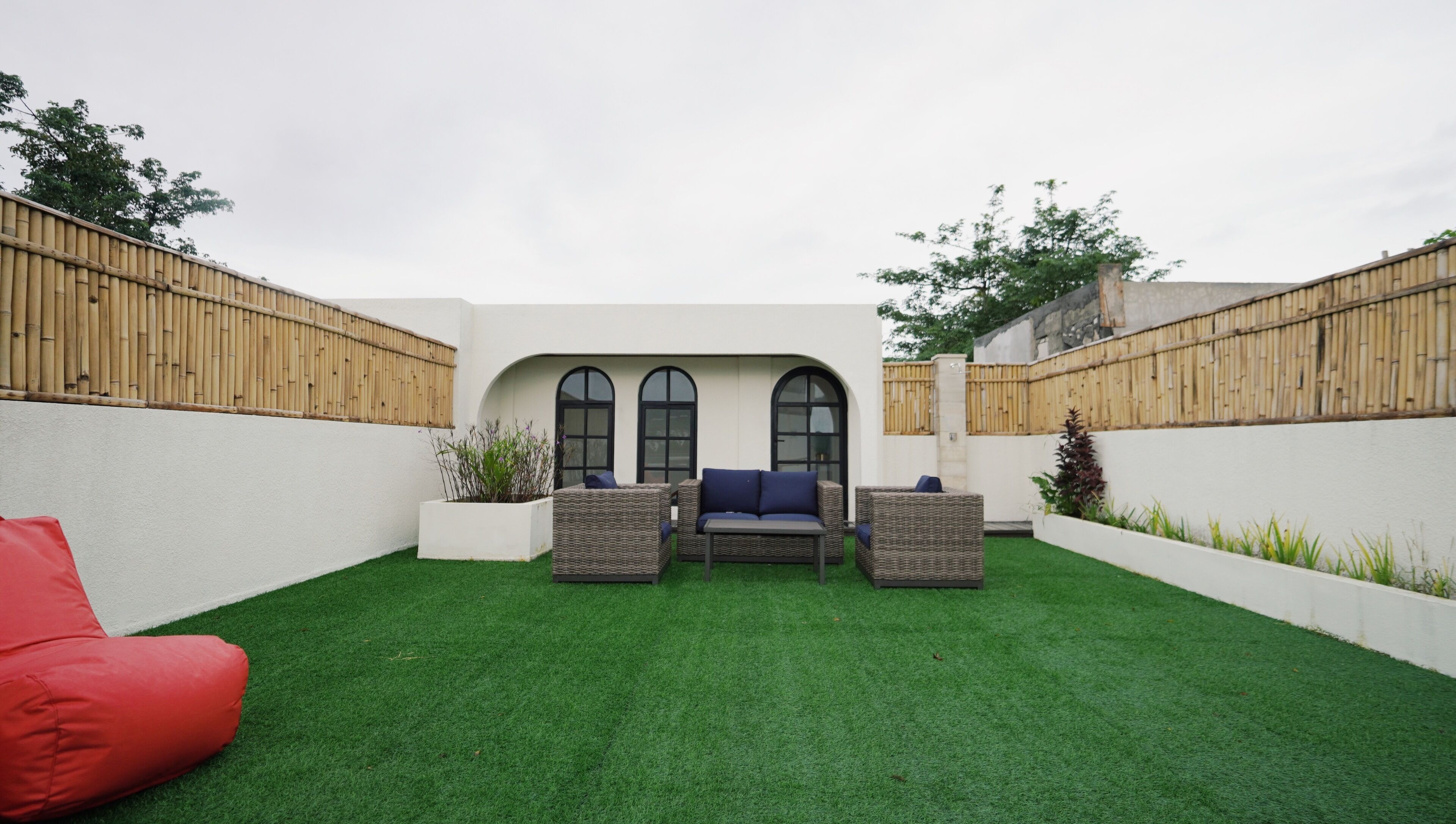 Un jardin avec une pelouse artificielle, un pouf rouge en forme de haricot, des meubles en osier et un mur blanc agrémenté de fenêtres en arc de cercle.