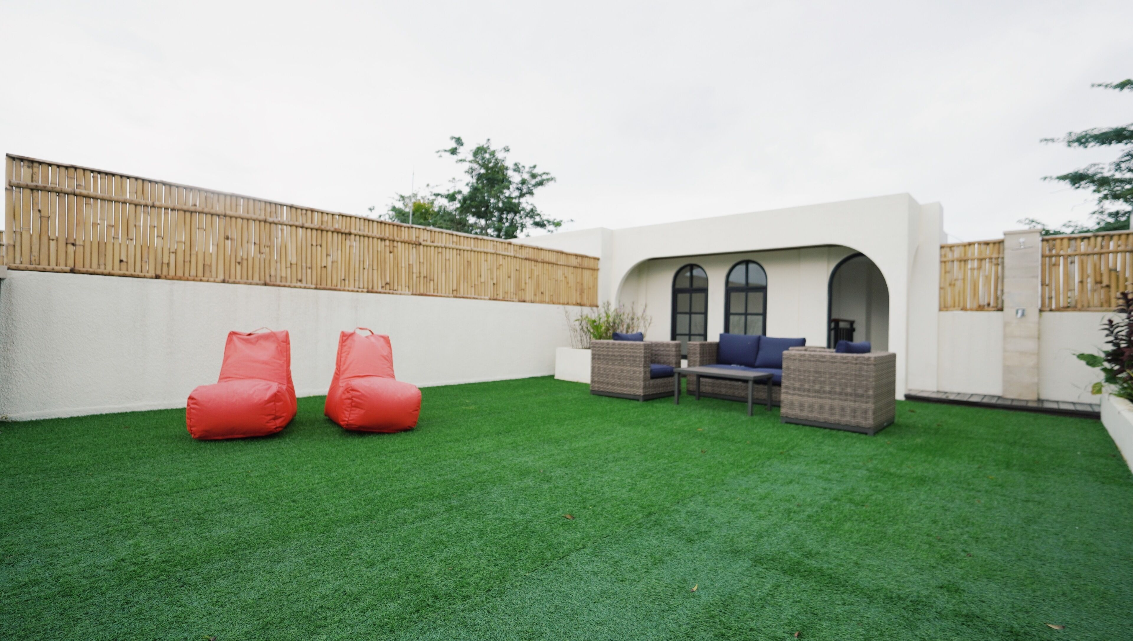 A backyard with artificial grass, a wooden fence, and outdoor furniture including two red bean bags and a wicker sofa with blue cushions.