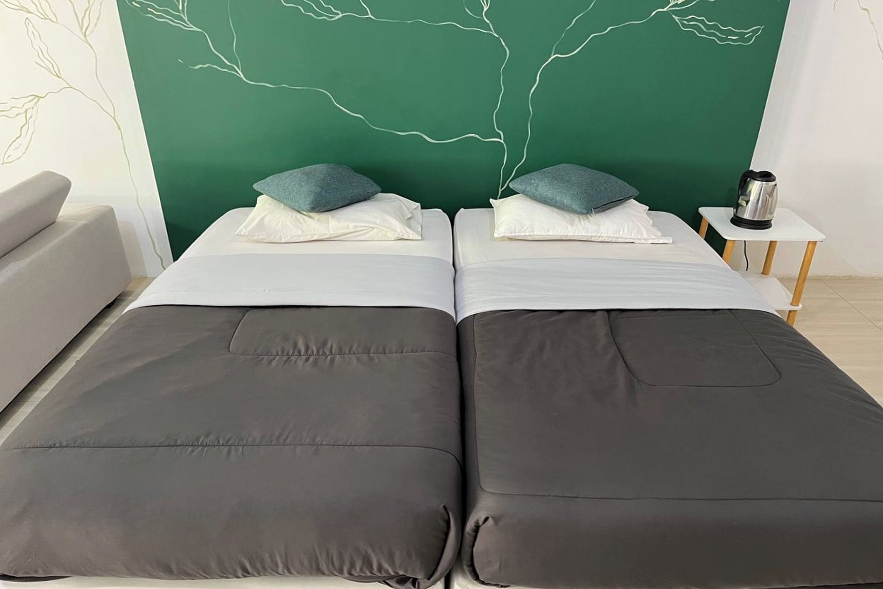 Une chambre avec deux lits, chacun doté de draps et d’oreillers gris, un mur vert orné d’un motif blanc en forme d’arbre, un canapé blanc et une petite table avec une bouilloire.
