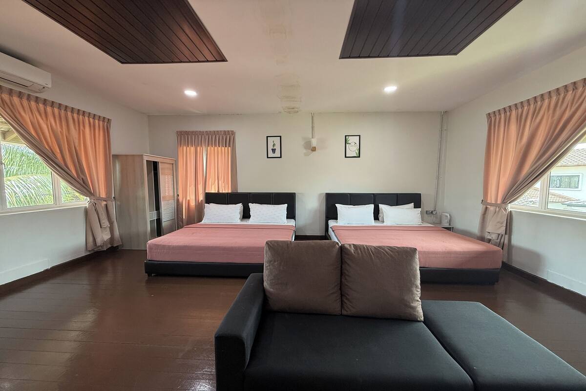 A hotel room with two beds, a sofa, wooden flooring, and large windows with curtains.
