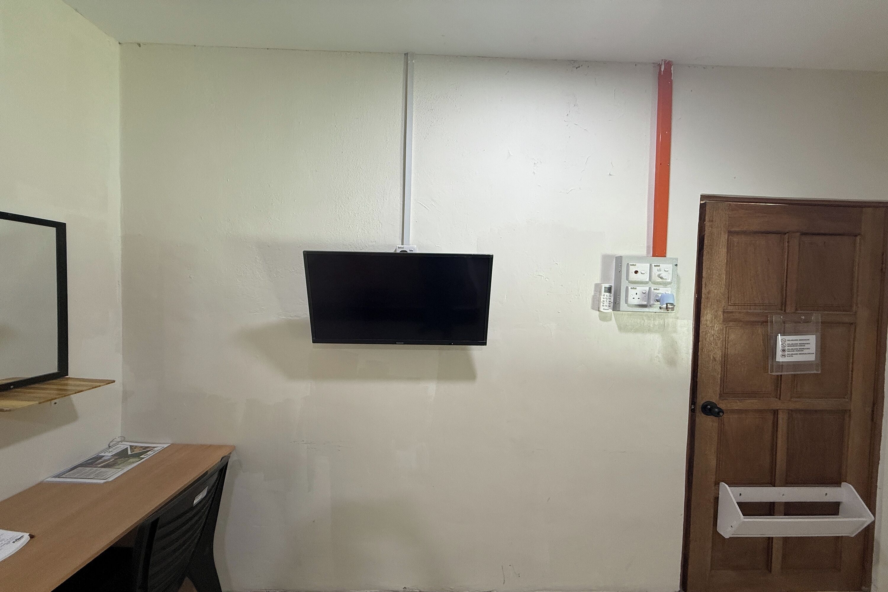 Una habitación con una televisión de pantalla plana montada en la pared, una puerta de madera con un buzón y un estante pequeño en la pared izquierda.
