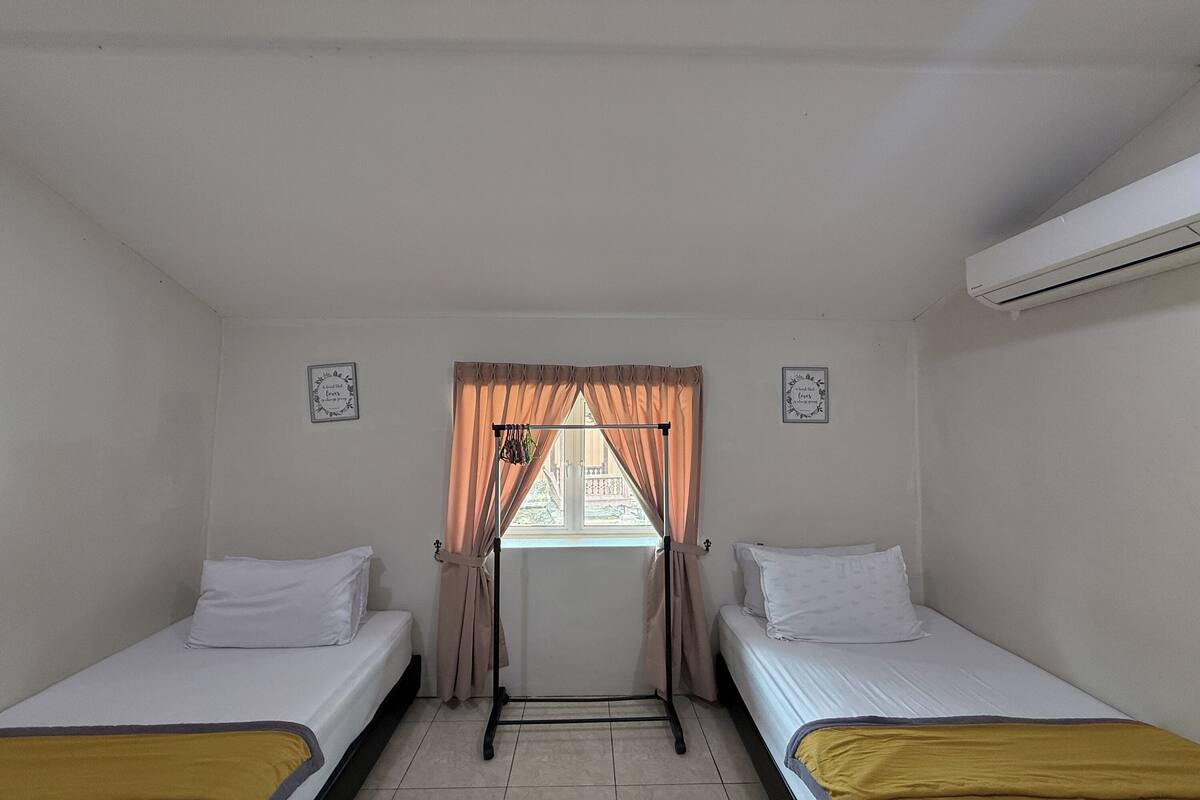 A small, clean room with two single beds, a window with curtains, and an air conditioning unit.