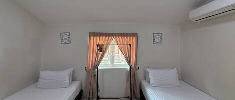 A small, clean room with two single beds, a window with curtains, and an air conditioning unit.