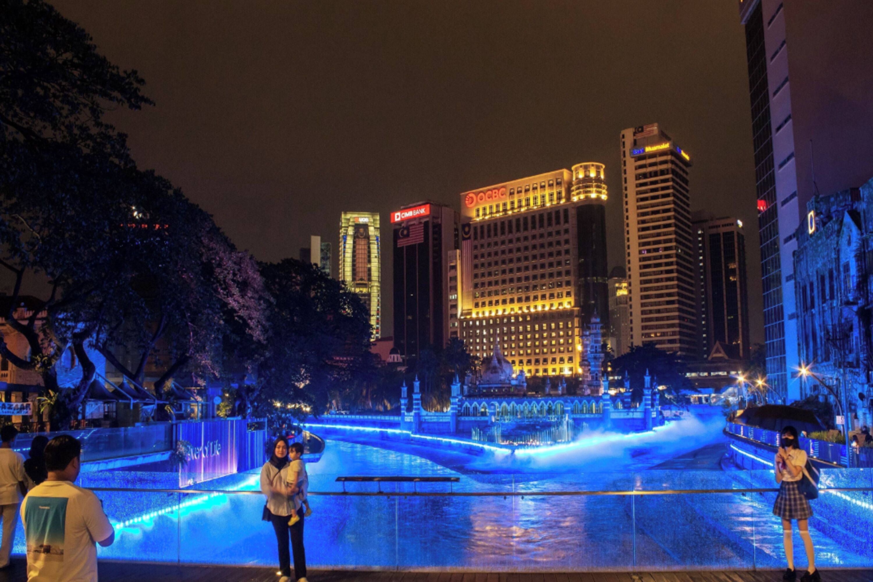 A nighttime cityscape with illuminated buildings and a water feature in the foreground.