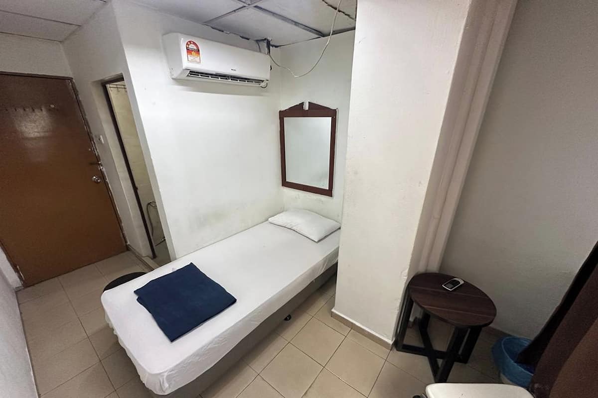Une petite chambre avec un lit simple, un matelas blanc, une couverture bleue, un miroir à cadre en bois, un petit tabouret et un appareil de climatisation fixé au mur.