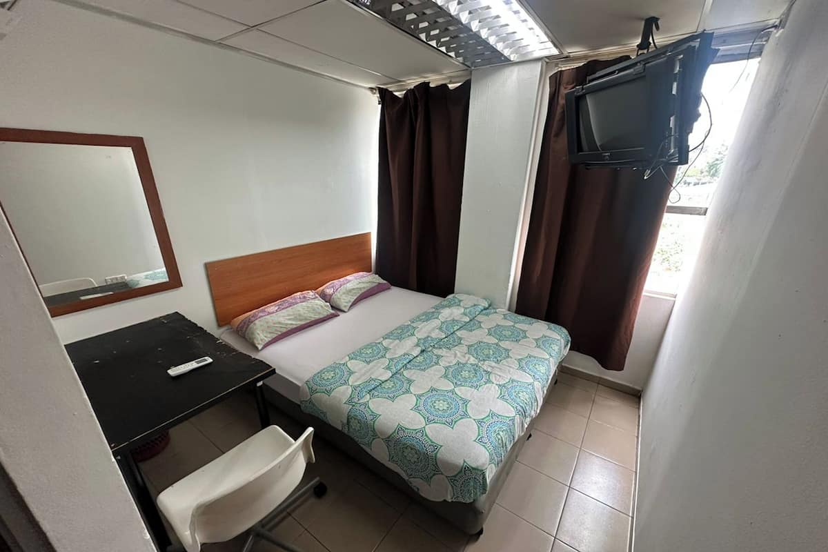Une petite chambre à coucher, bien rangée, avec un lit, un bureau, une chaise, un téléviseur fixé au mur et une fenêtre avec des rideaux.