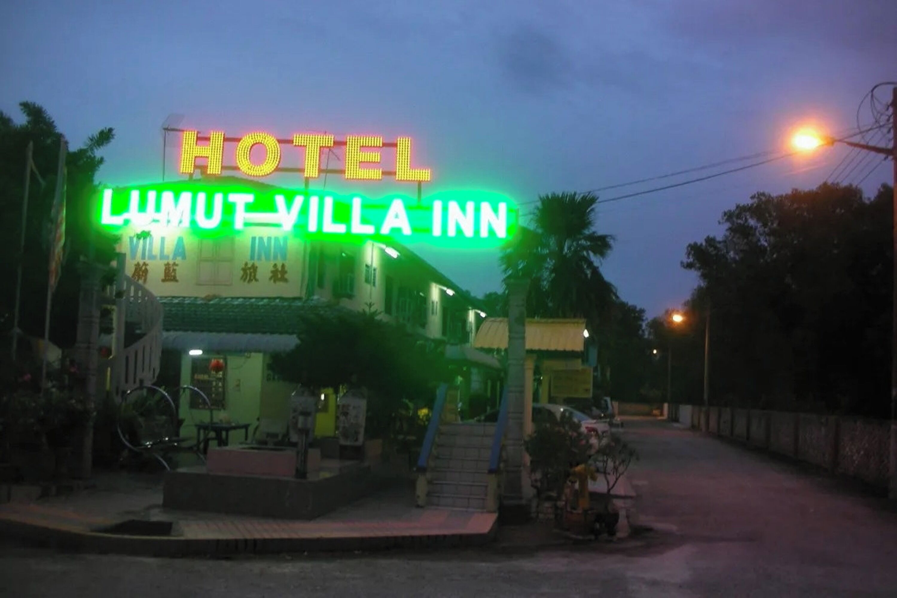 A hotel named "LUMUT VILLA INN" with a prominent neon sign, a staircase leading to the entrance, and a small outdoor seating area with tables and chairs.