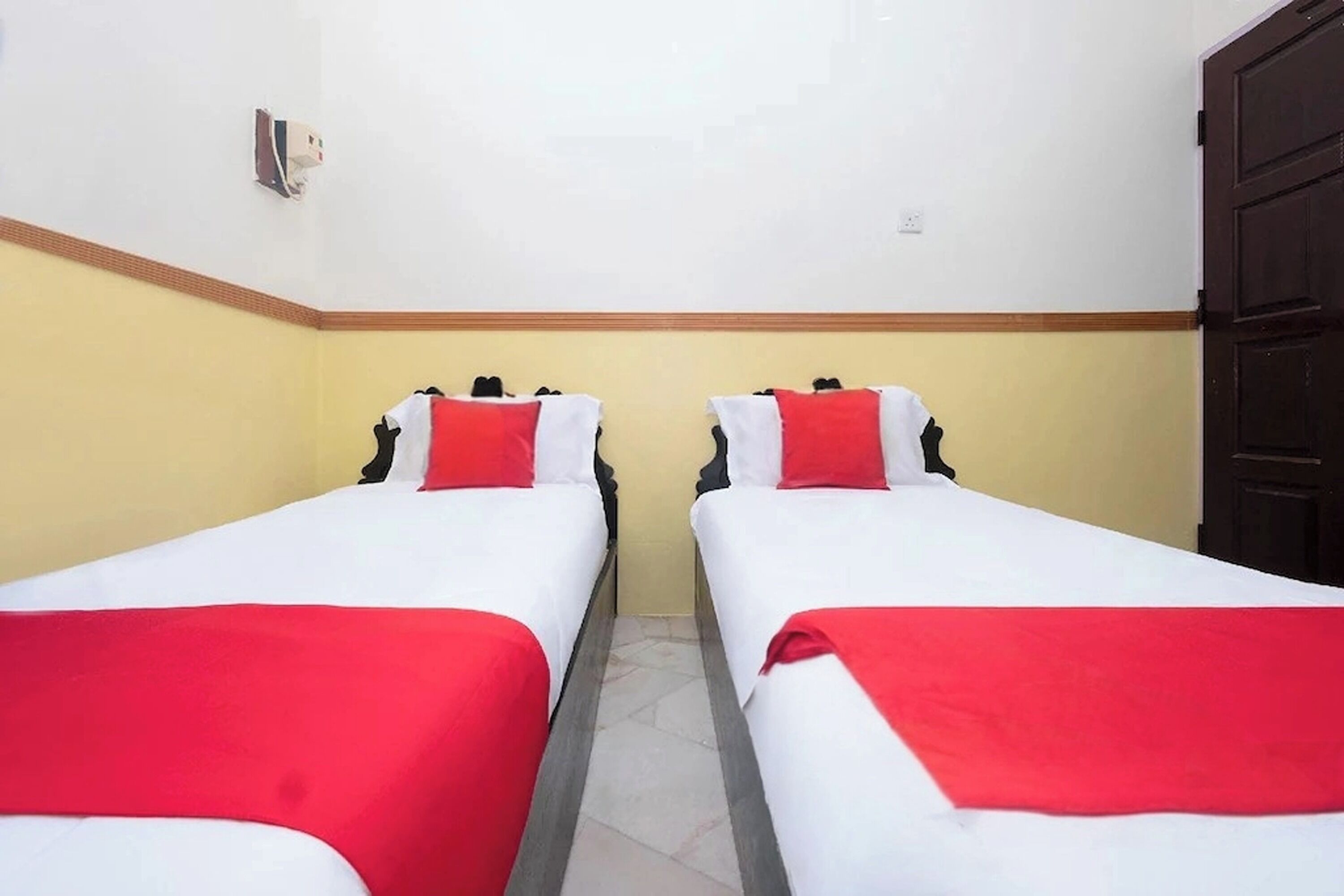 Een hotelkamer met twee éénpersoonsbedden, elk met een rood-witte spreidek en kussens. De kamer heeft een houten deur en een lichtgekleurde wand met een gele rand.