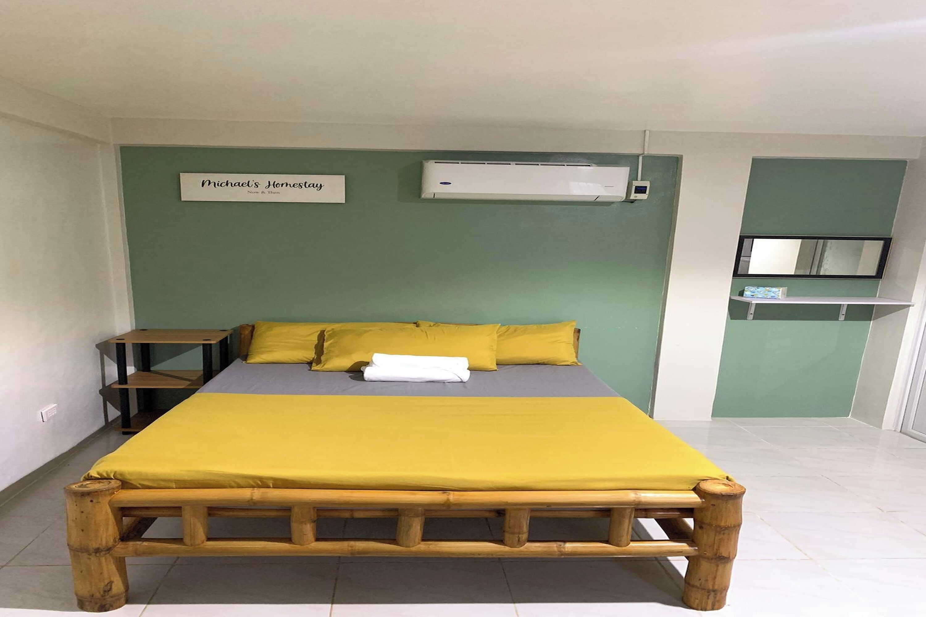 Habitación individual con cama de madera, colcha amarilla y colchón gris. Cuenta con una mesita de noche con lámpara, aire acondicionado de pared y un cartel sobre la cama.