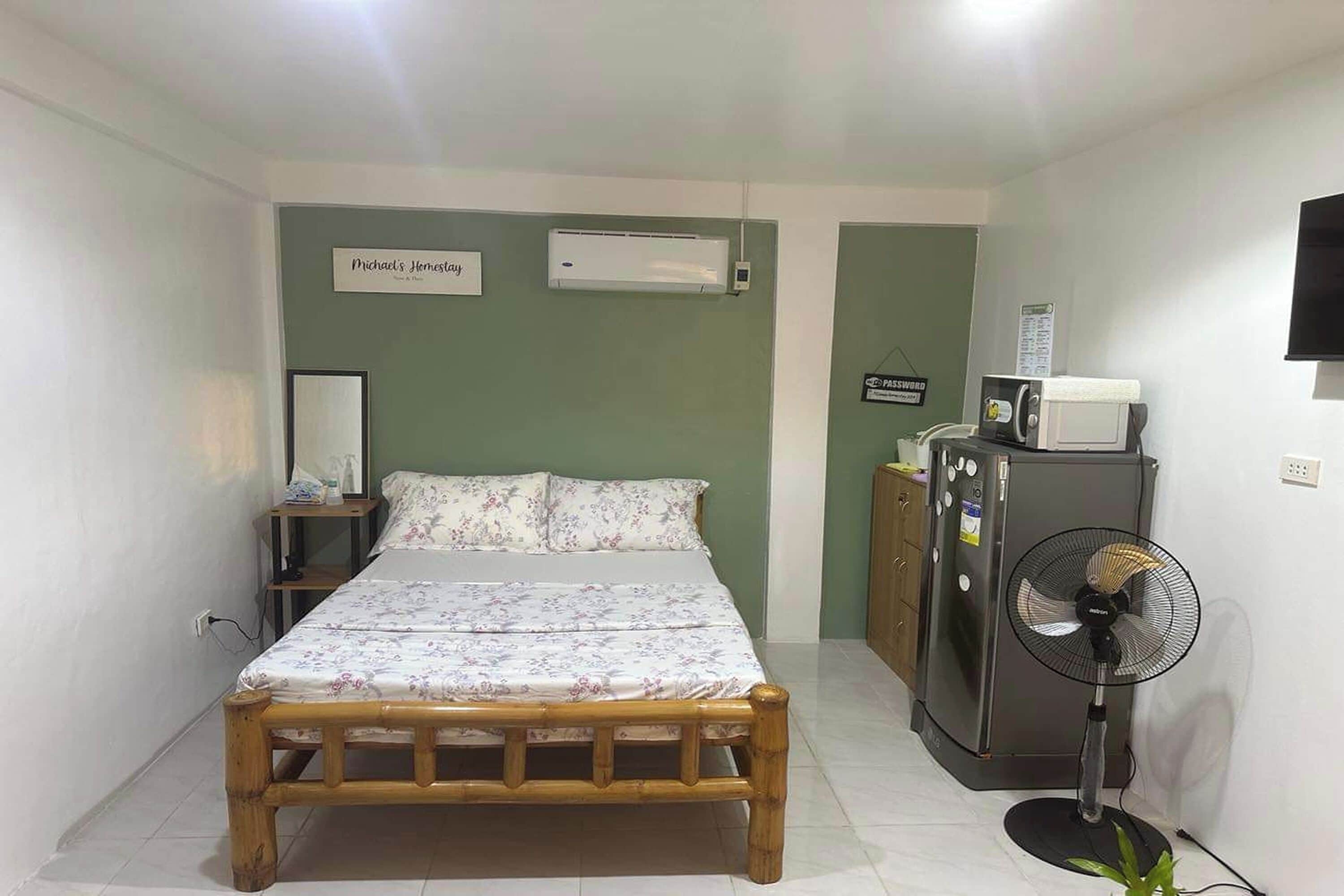 Une chambre comprenant un lit, une table de chevet, un climatiseur, un réfrigérateur, un four à micro-ondes et un ventilateur sur pied.