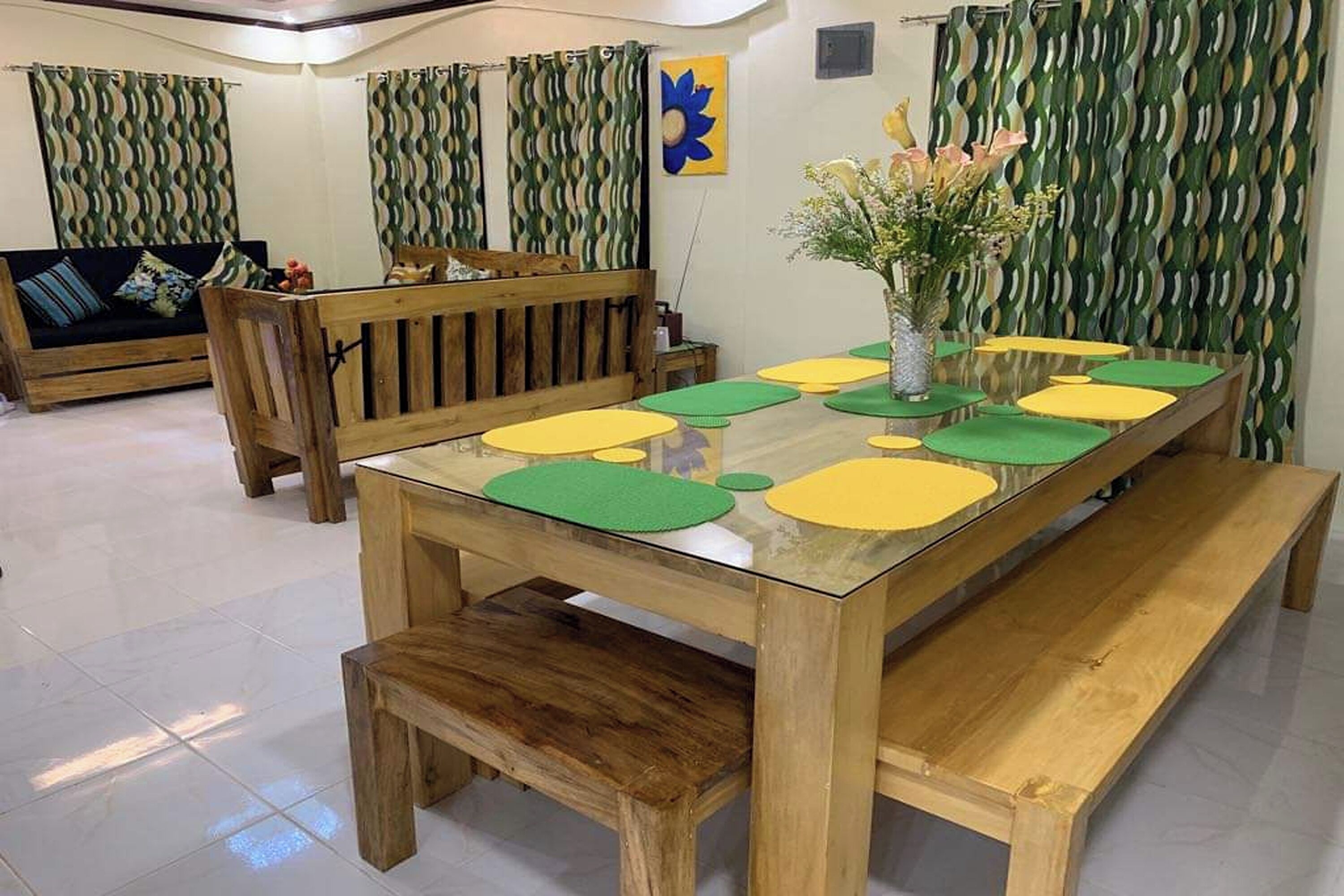 Un comedor con una mesa de madera y bancos, un jarrón con flores y cortinas estampadas.