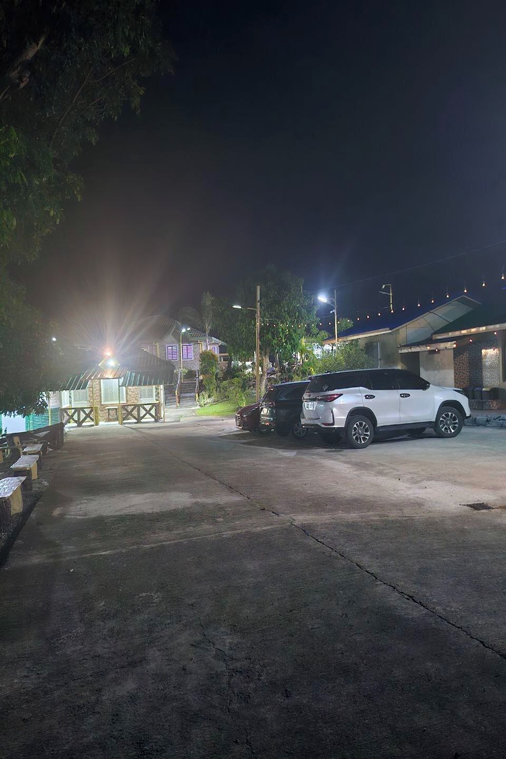 A parking area with two cars, a building with a sloped roof, and a well-lit pathway leading to a gated entrance.