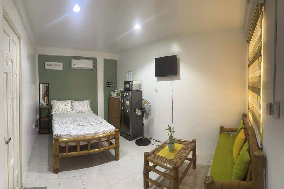 Ein Schlafzimmer mit Bett, Nachttisch, Ventilator, Fernseher und einem kleinen Tisch mit einer Pflanze.