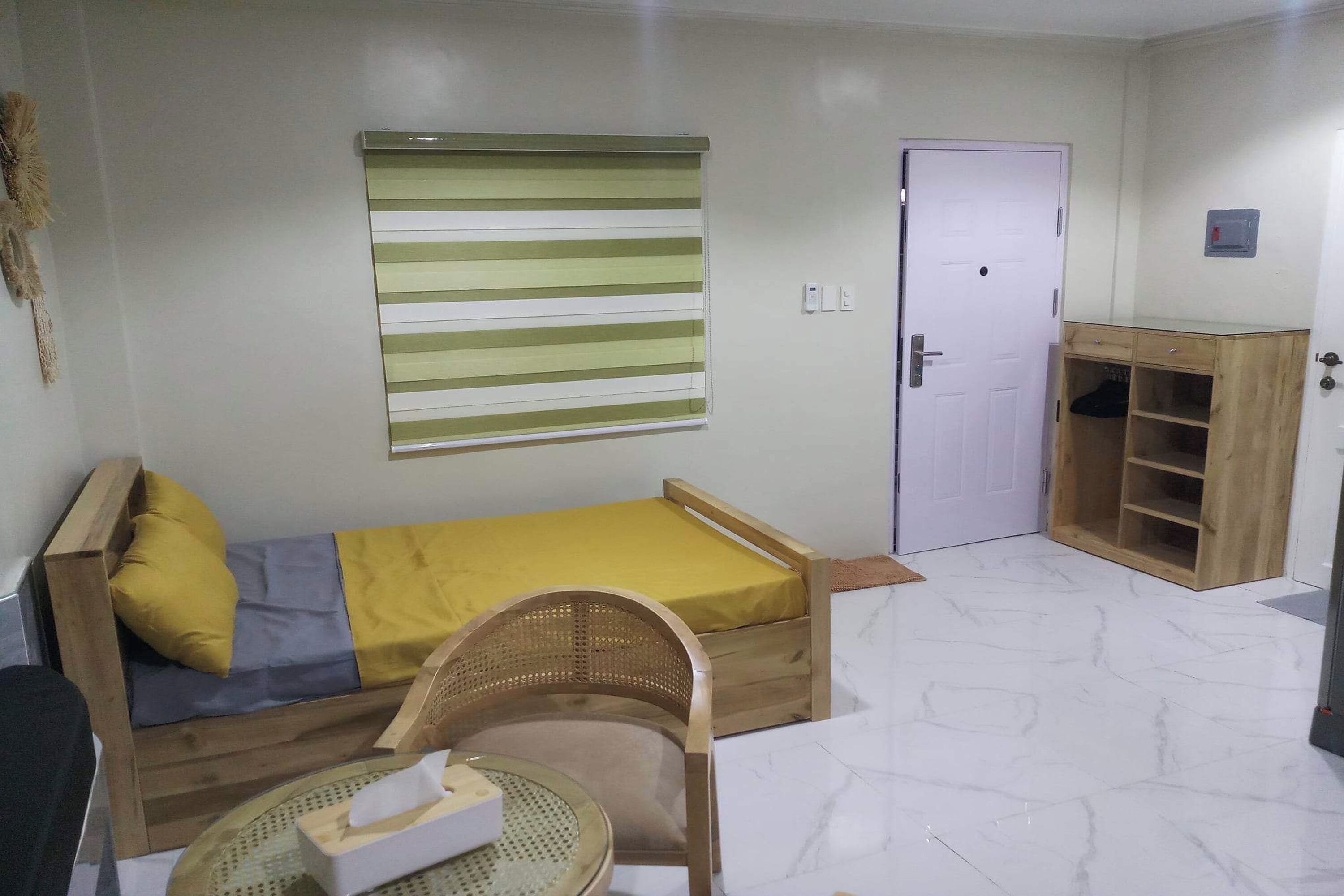 A small, furnished room with a single bed, a wooden cabinet, a door, and a window with blinds.