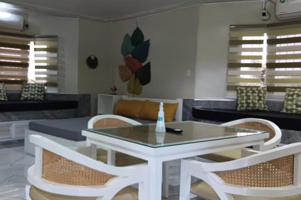 Uma sala de estar moderna com uma mesa de vidro, cadeiras de vime branco, um sofá com almofadas e um ar condicionado fixado na parede.
