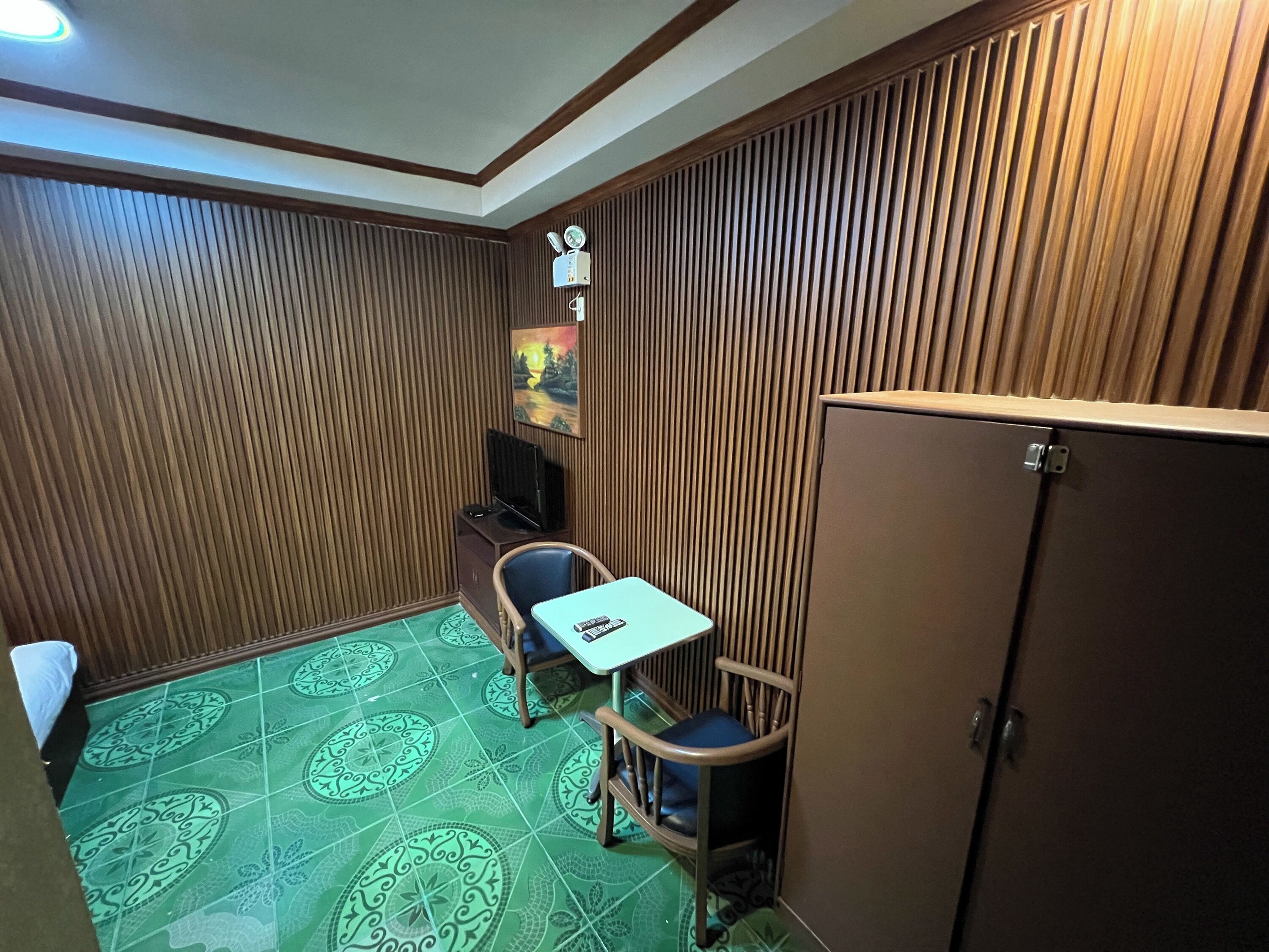 Una habitación pequeña con un muro de listones de madera, una mesita con una silla, un armario y un televisor sobre un soporte.