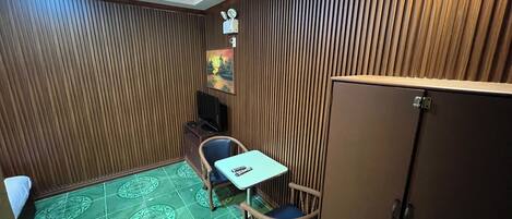 Una habitación pequeña con un muro de listones de madera, una mesita con una silla, un armario y un televisor sobre un soporte.