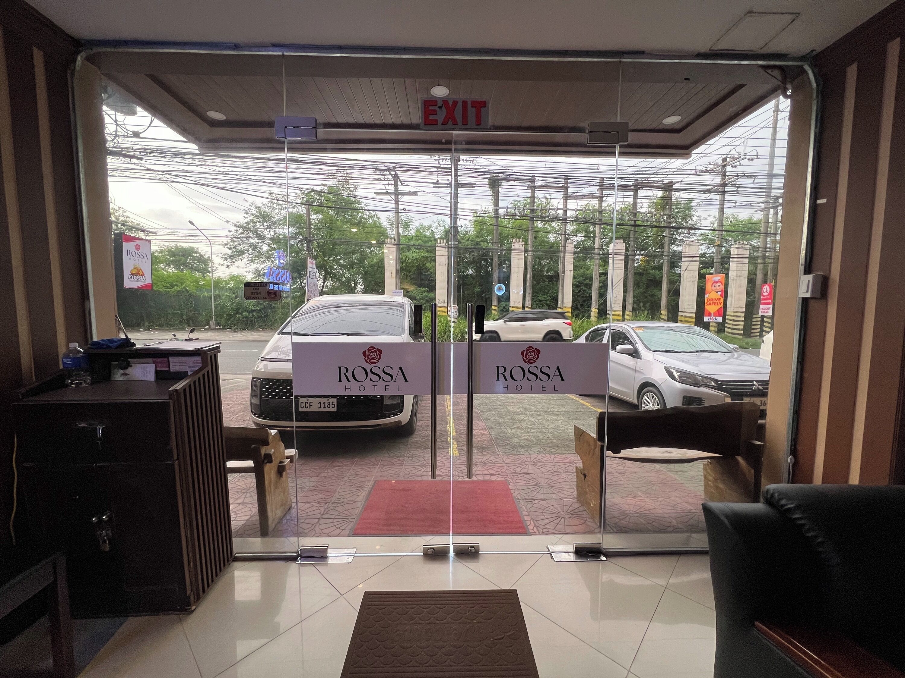 A hotel entrance with glass doors, a reception desk, and a view of parked cars outside.
