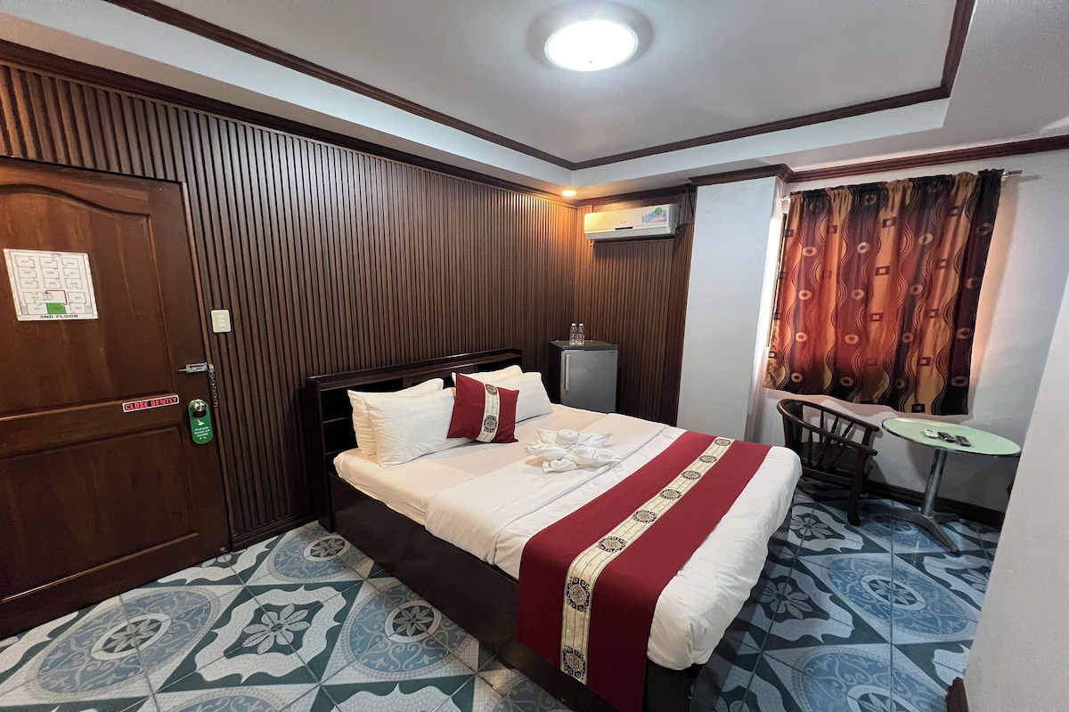 A hotel room with a wooden panel wall, a bed with white bedding and a red blanket, a small table, and a chair.