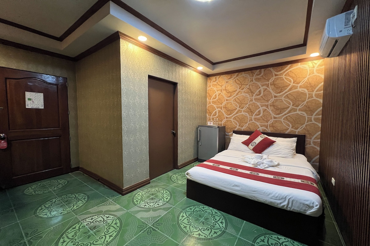 A hotel room with a bed, a wardrobe, a small cabinet, and patterned green floor tiles.