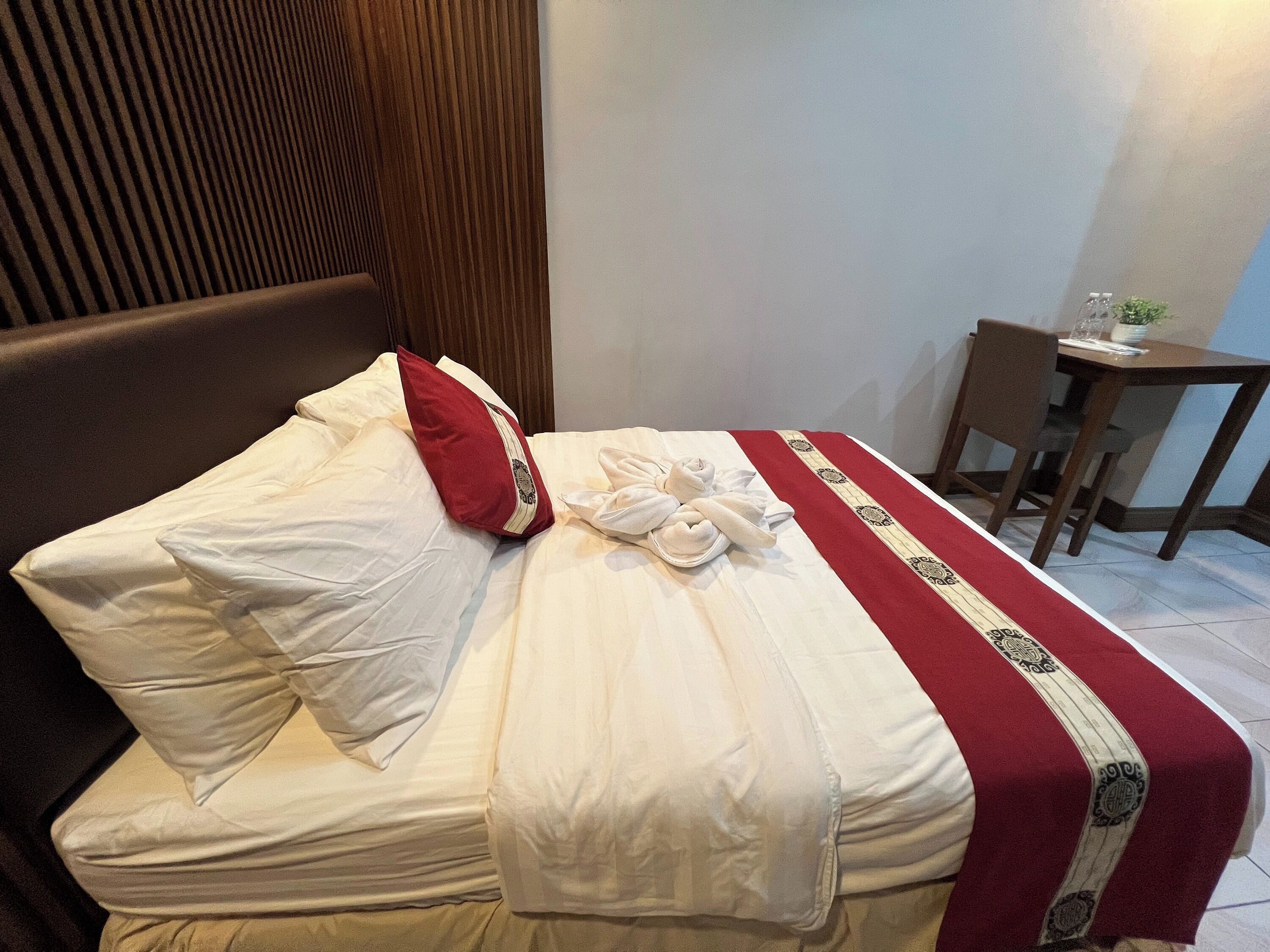 Una cama bien hecha con sábanas blancas, un cojín rojo y una colcha decorativa roja y blanca. En la esquina, hay una mesita con una planta y una silla.