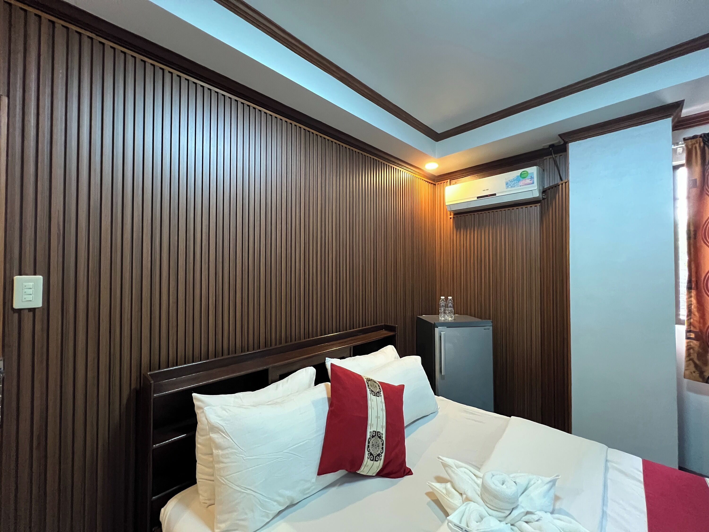 A hotel room with a bed, a nightstand, a minibar, and a ceiling light.