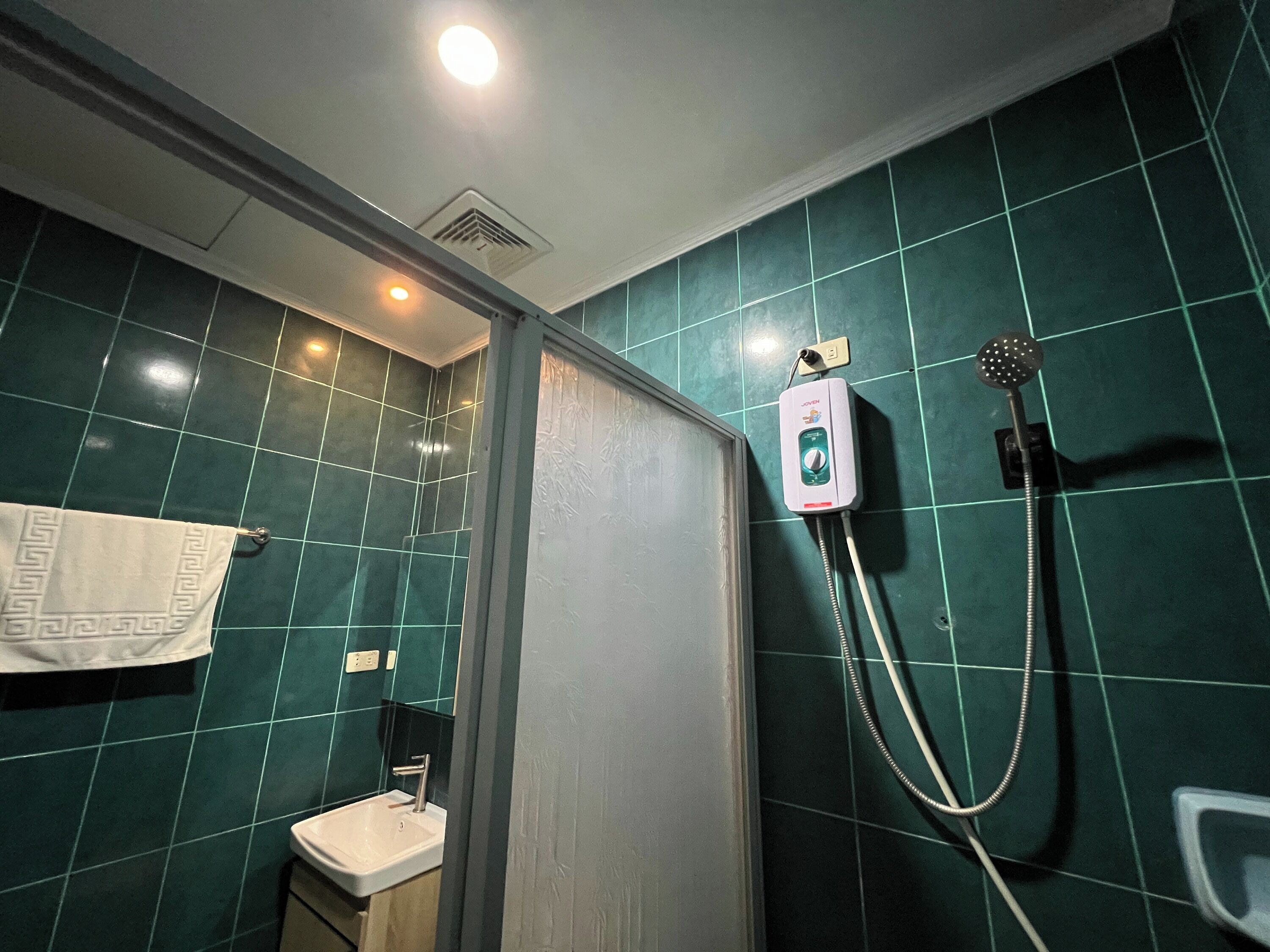 Un baño con baldosas verdes, un lavabo blanco, una toalla y una ducha con cabezal de ducha manual.