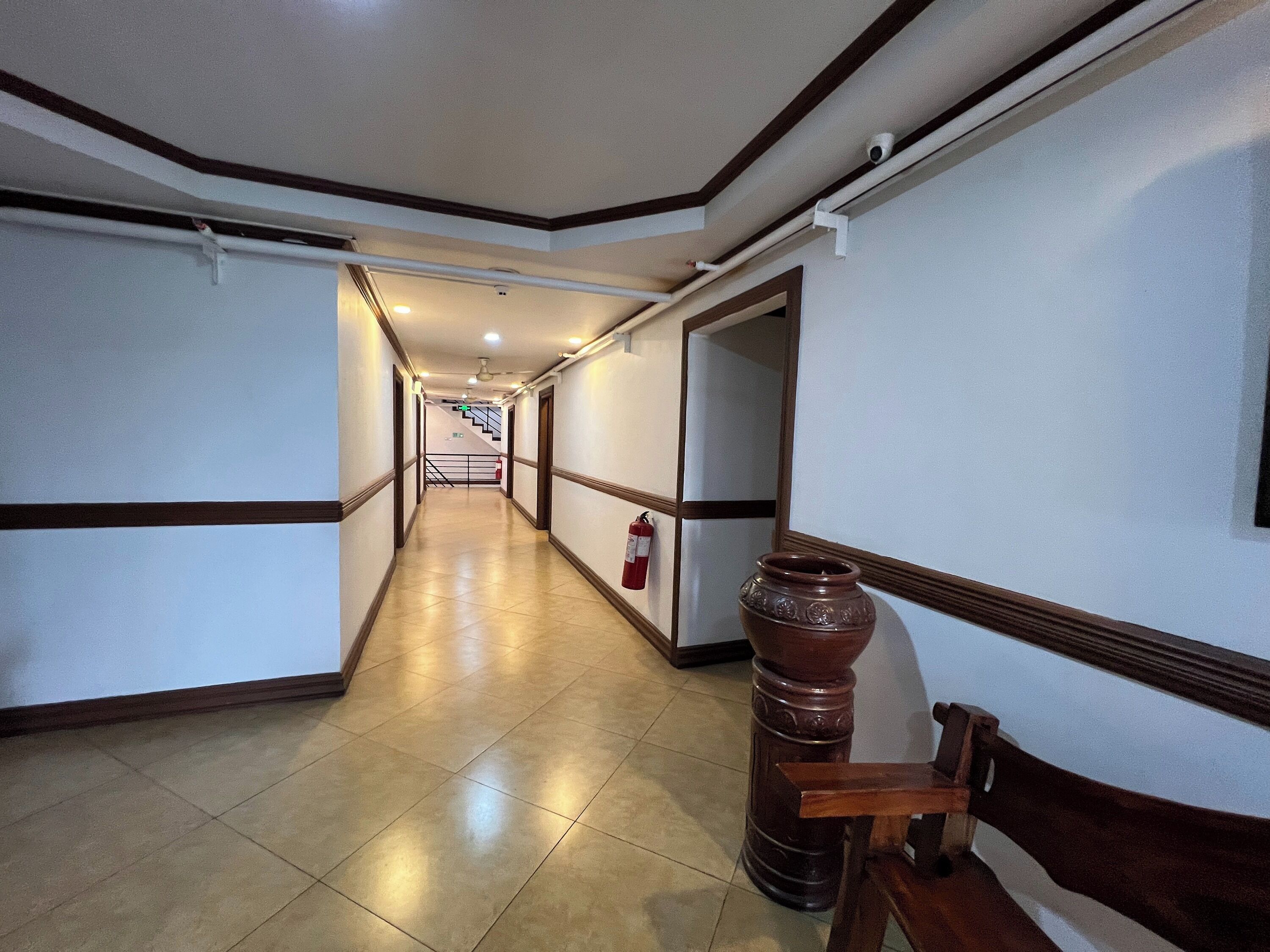 A hallway with a tiled floor, a wooden bench, a large vase, and a fire extinguisher.