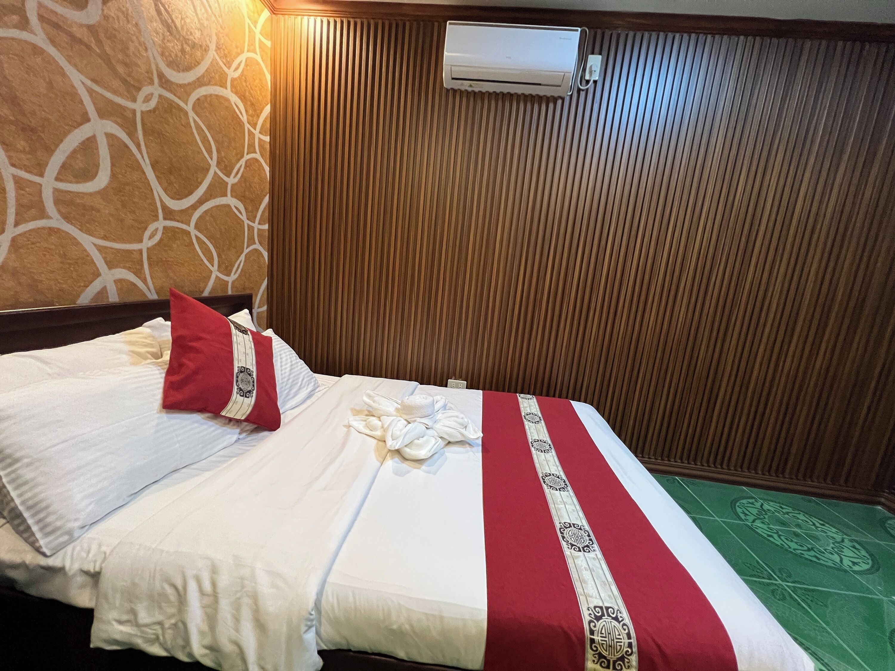A hotel room with a bed, a red pillow, a white pillow, a red and white bedspread, and an air conditioning unit on the wall.