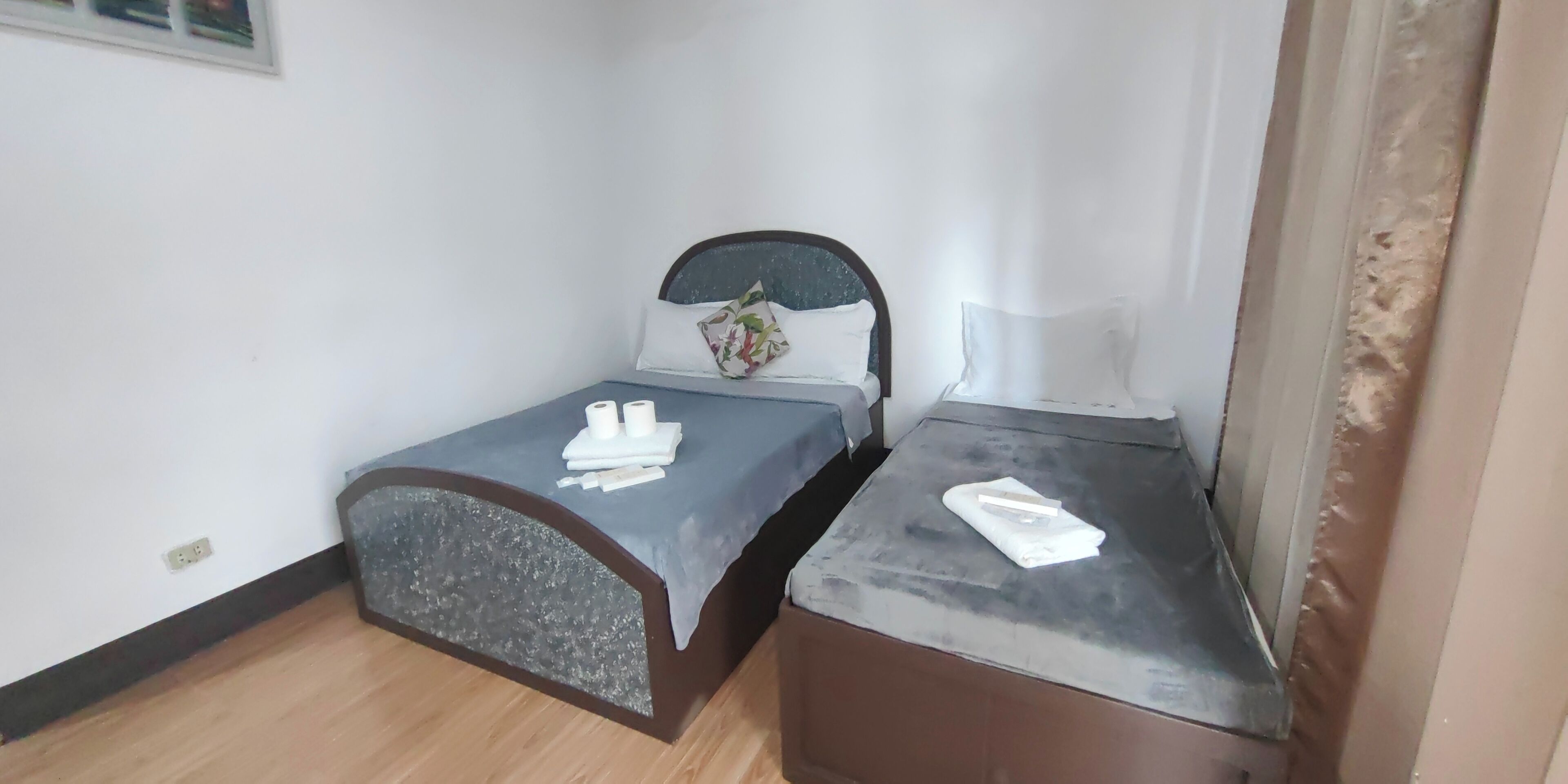 Une petite chambre avec un lit simple, un plancher en bois, une tête de lit agrémentée d’un coussin à motifs floraux et une table de chevet avec une lampe.