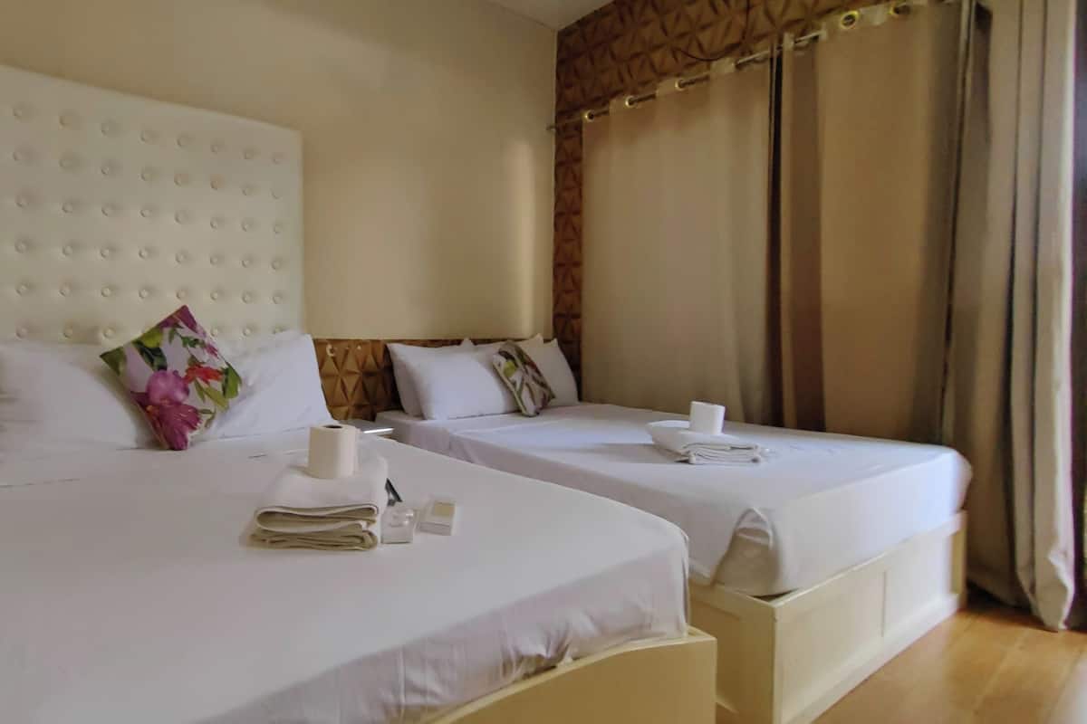 A hotel room with two beds, each with a white headboard and pillows. There are toiletries on the beds, and a curtain is partially drawn to the side.