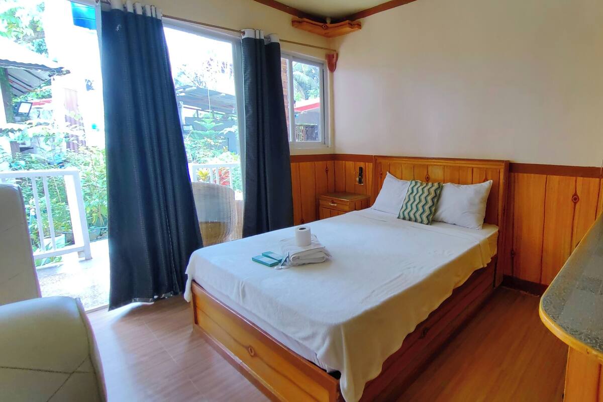 A bedroom with a wooden bed, a small bedside table, a window with curtains, and a view of greenery outside.