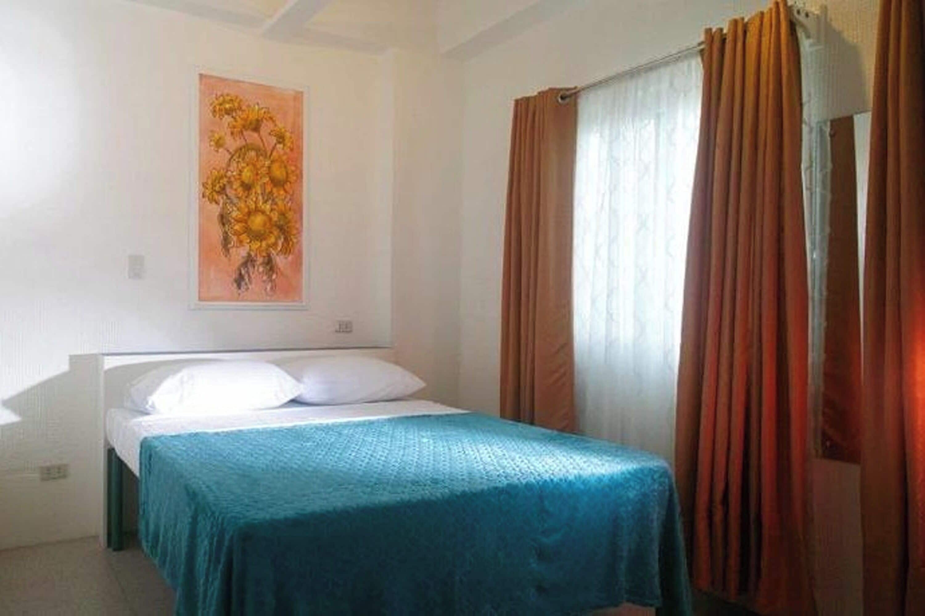A single bed with a teal bedspread, a painting of flowers on the wall, and a window with curtains.