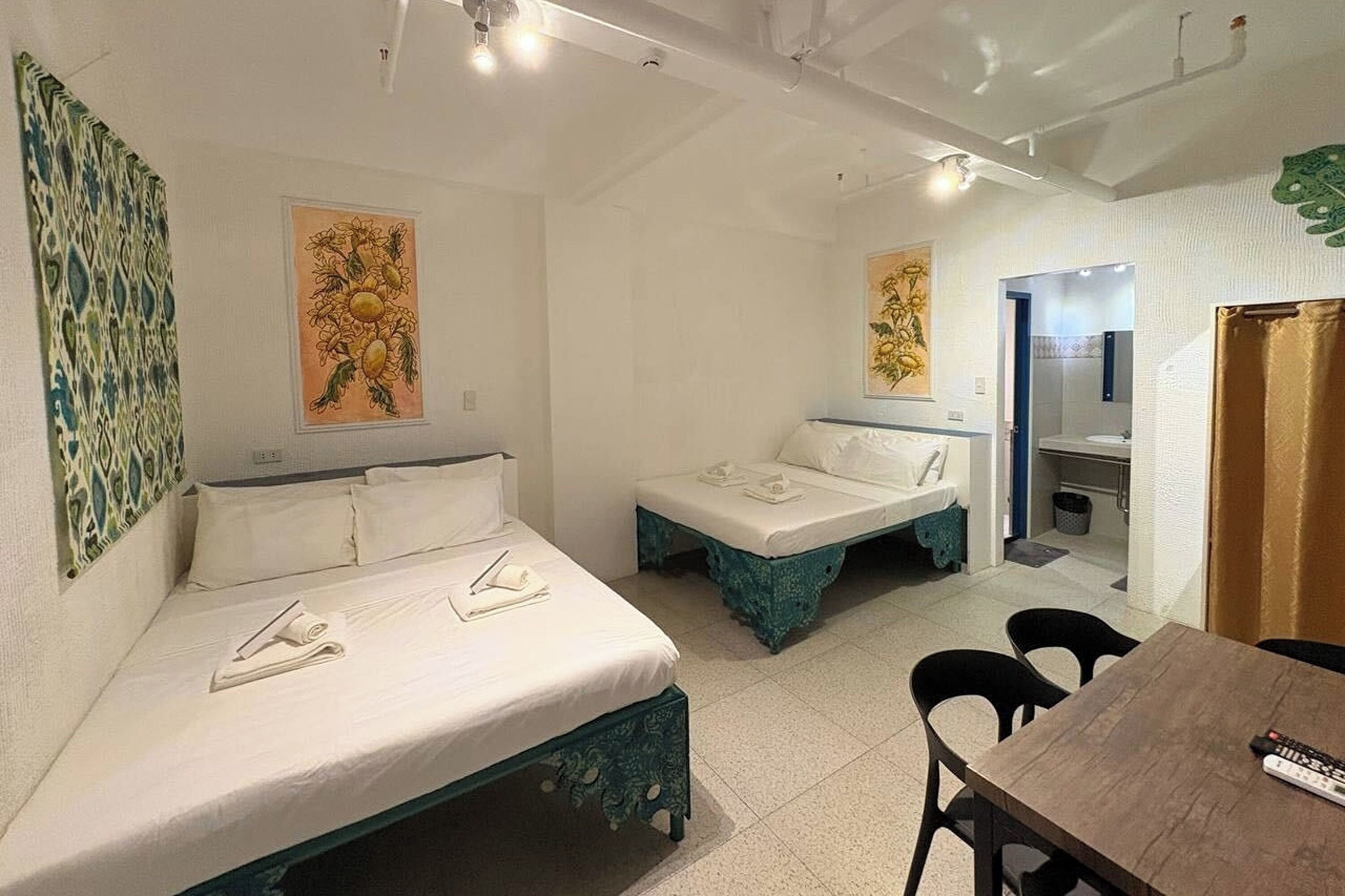 A room with two beds, a table, and a chair. There is a bathroom visible through an open door.