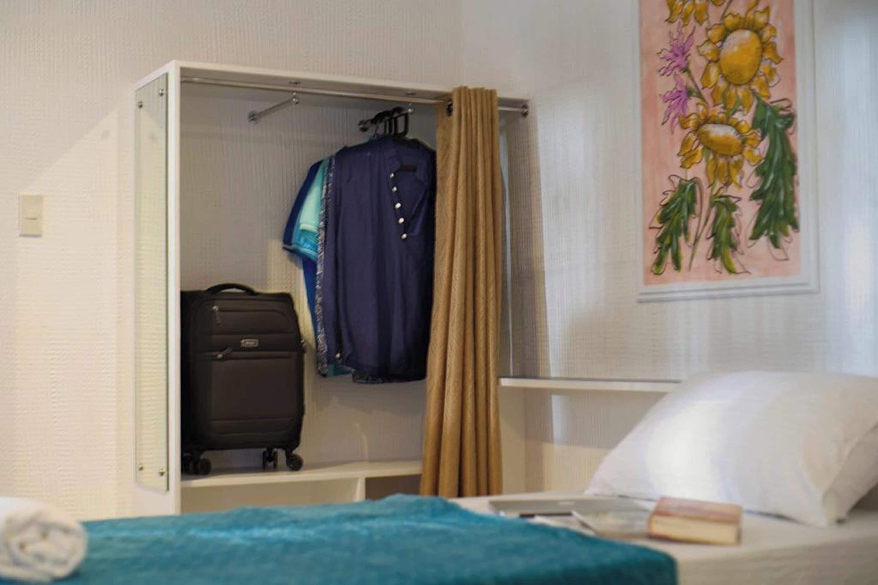 A room with a bed, a suitcase, a hanging coat, and a painting of flowers.