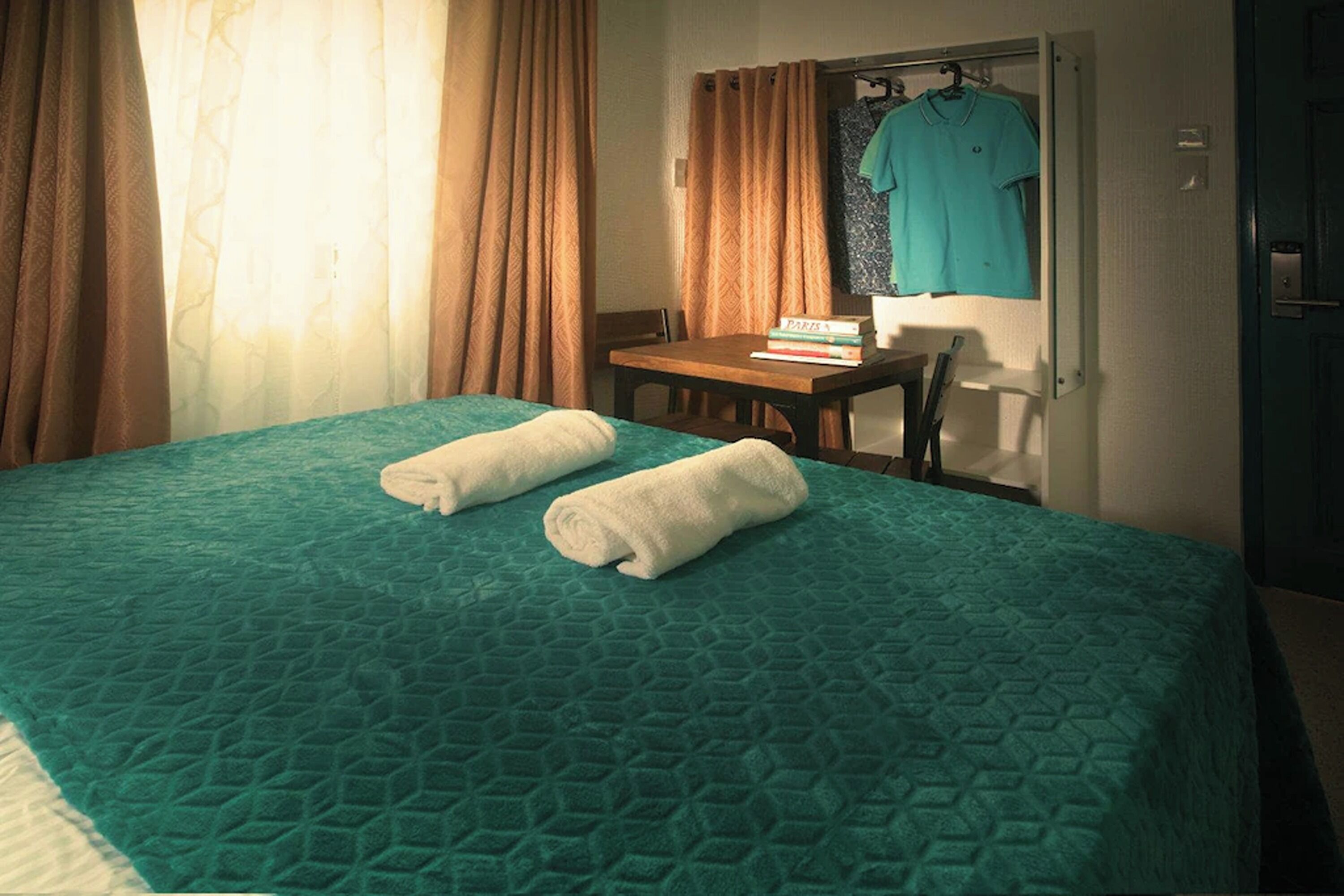 Una habitación de hotel con una cama, una mesa con libros, una camisa colgada y una puerta.