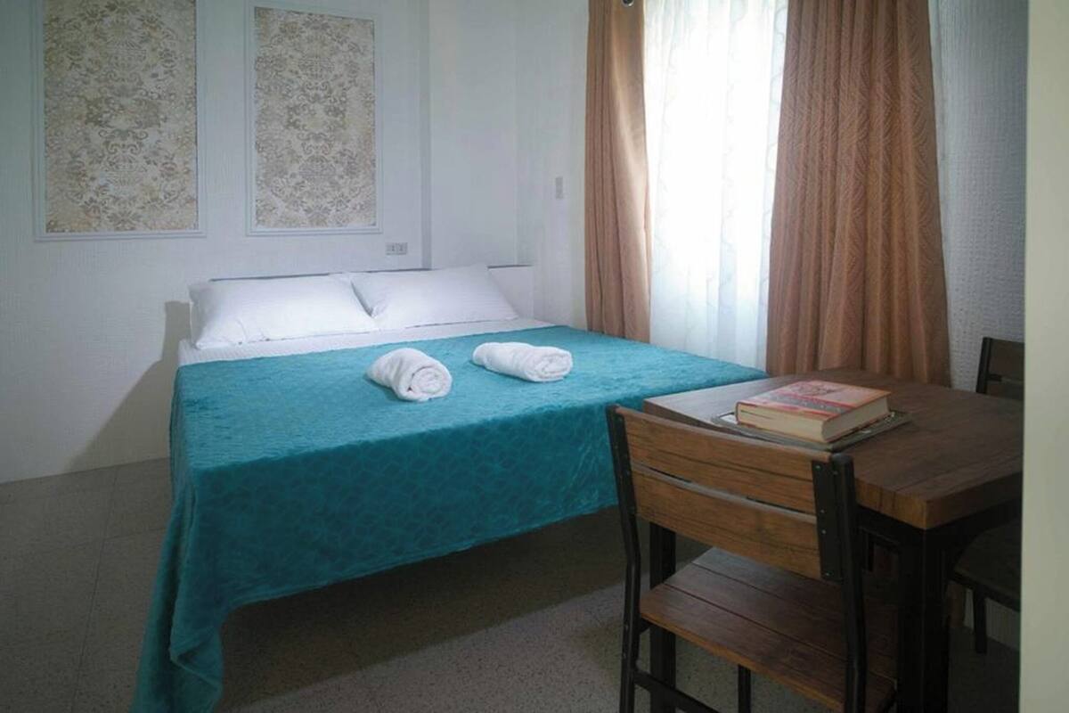 Un letto singolo con una coperta color petrolio, due asciugamani arrotolati e un libro su un tavolo di legno.
