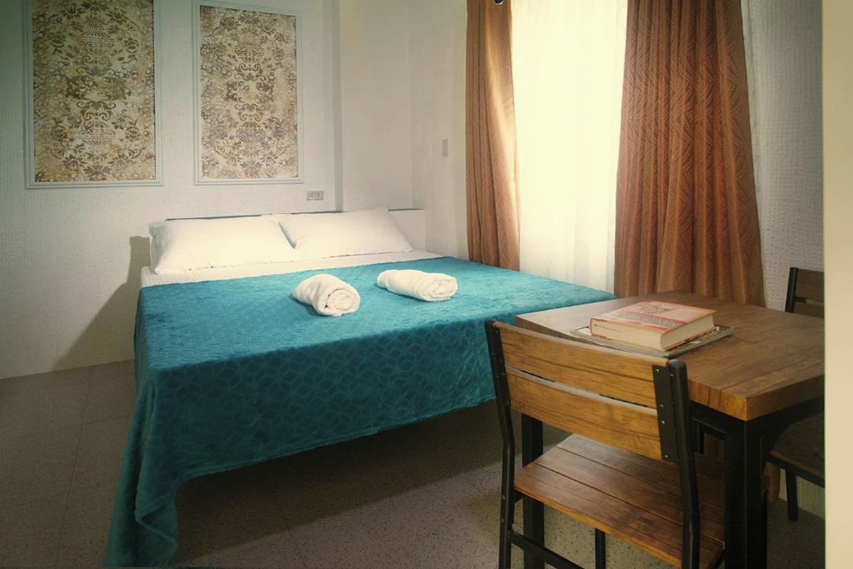 Un letto singolo con copriletto color petrolio, una sedia di legno, un tavolo con un libro e due decorazioni murali a motivi